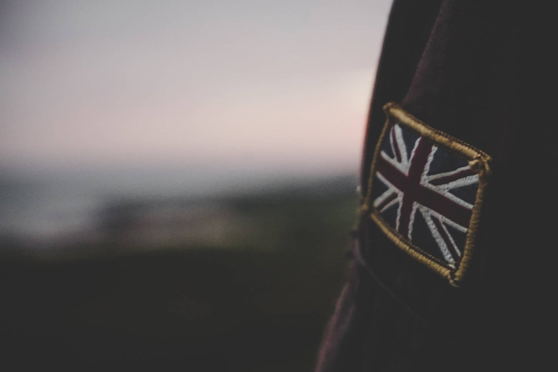 A close up of the left arm of a Barbour wax jacket showing a Union Jack patch with a country scene blurred in the background