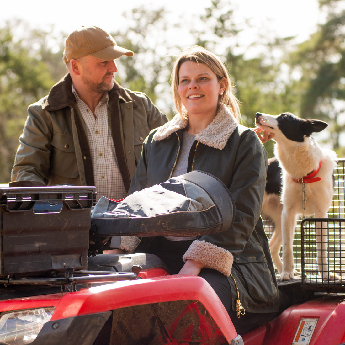 A man and woman sit on a tractor wearing Barbour and GANT wax jackets. Their dog sits behind