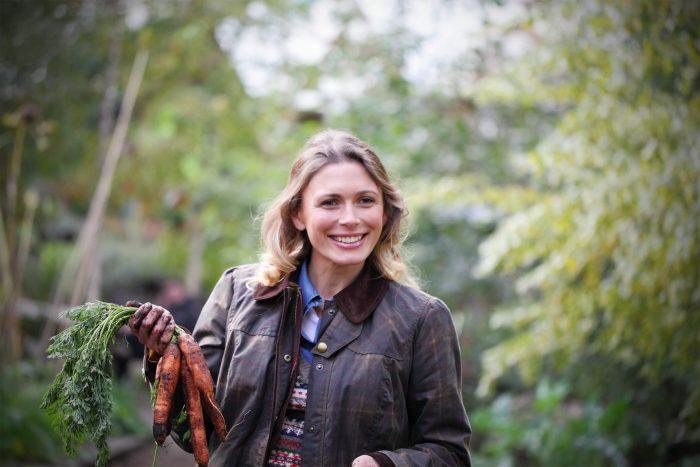 White woman with blonde hair wears tartan Barbour wax jacket and is holding carrots in the woods