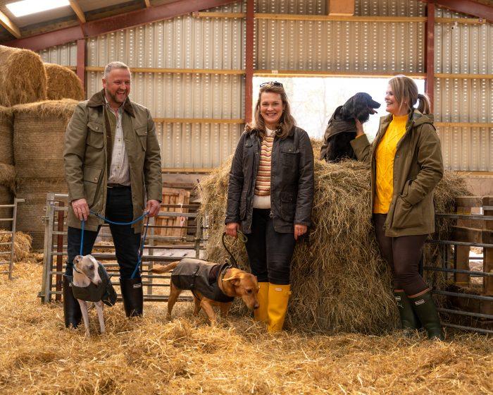 man and two women with dogs in a barn wearing fashionable country clothing