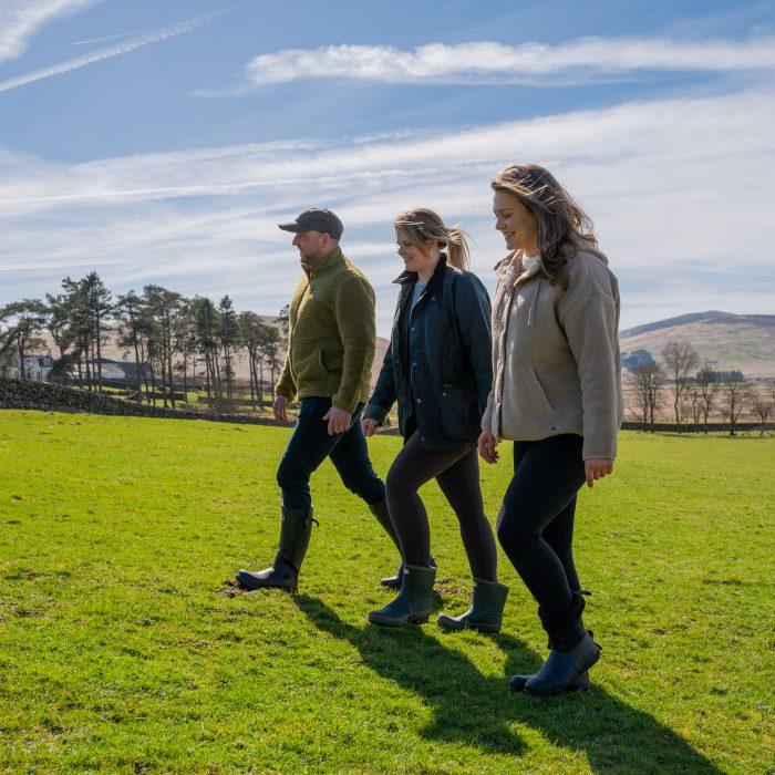 a man and two women walk through a grass field. The sun is shining and they are dressed in stylish countryside clothing