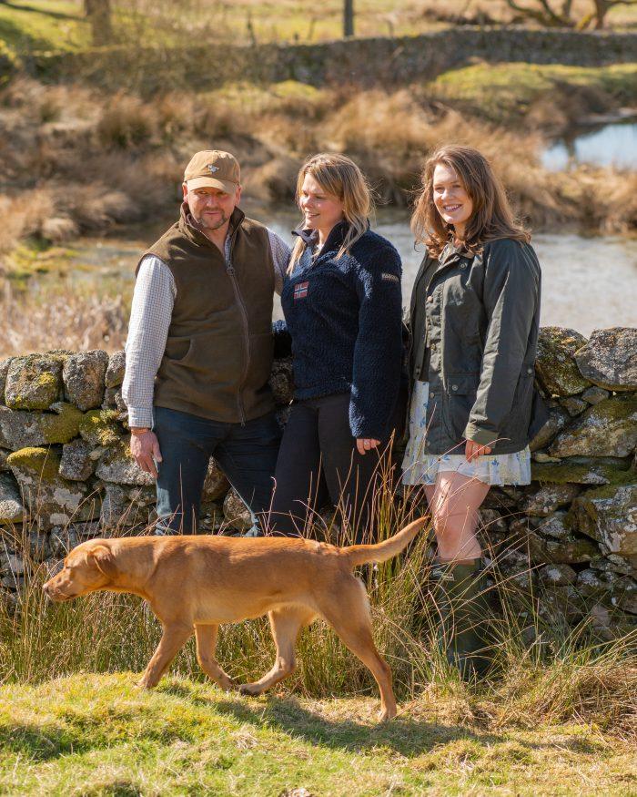 man and two women outside stand against a stone wall wearing fashionable country clothing. They have a golden retriever with them