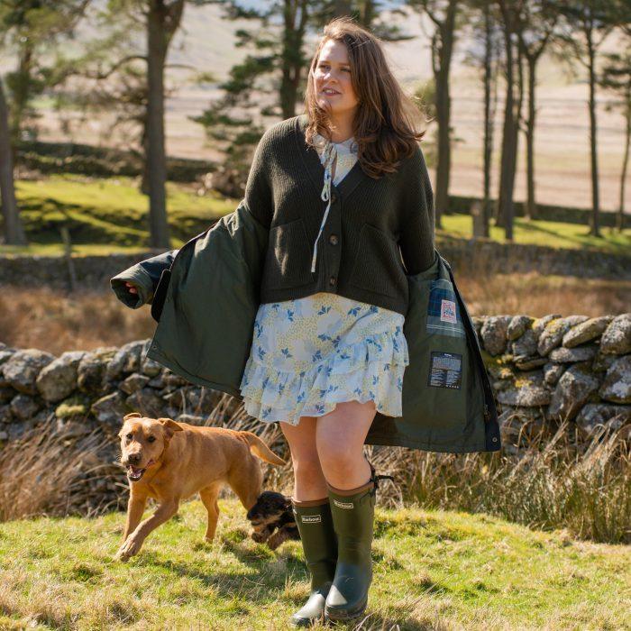 woman with dog explores the countryside in a floral dress, cropped cardigan, Barbour wellies and wax jacket