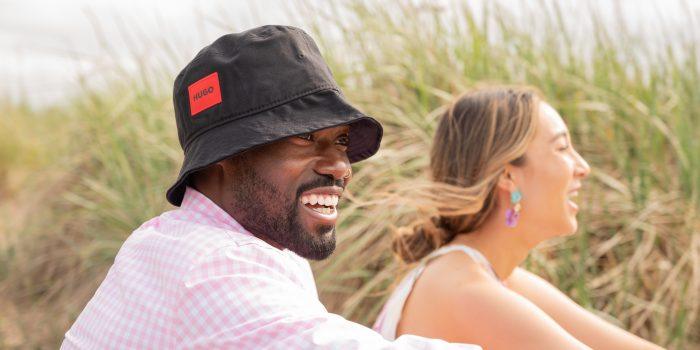 Man wearing a bucket hat sits with a woman laughing on the beach