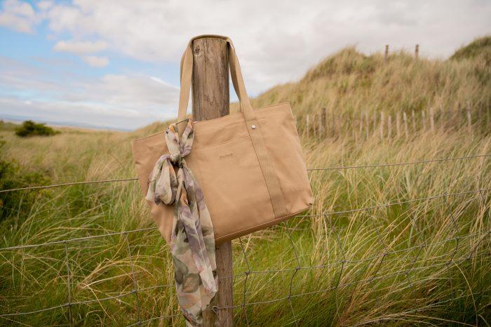 A beige Barbour bag hangs on a post at the beach. It has a flowered scarf attached to it