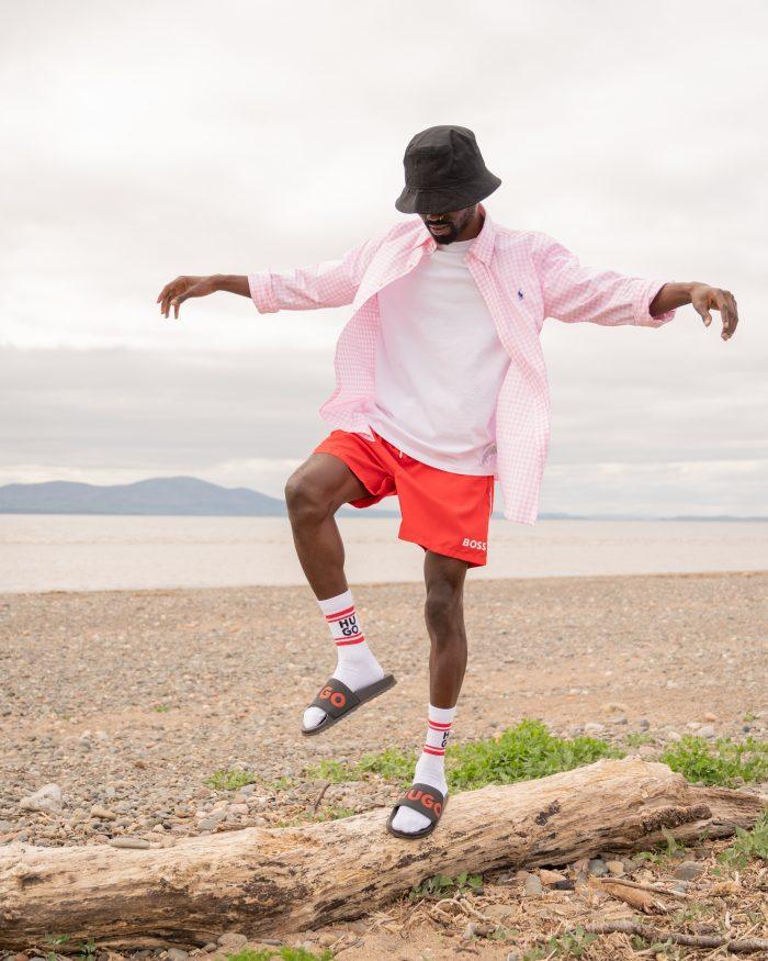 Man wearing shorts, a hat and an open shirt balances on a log at the beach