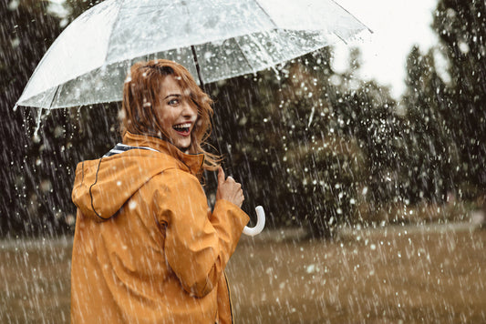 A woman stands under a clear umbrella in a mustard coloured raincoat. It's raining and she's smiling