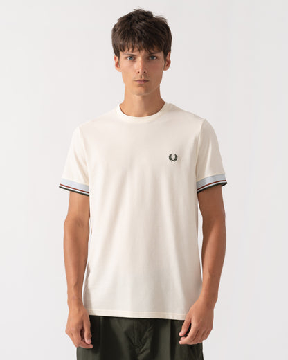 A man wearing a cream-colored short-sleeve t-shirt with a small embroidered logo on the left chest and striped ringer cuffs, paired with dark green pants, against a plain light gray background.