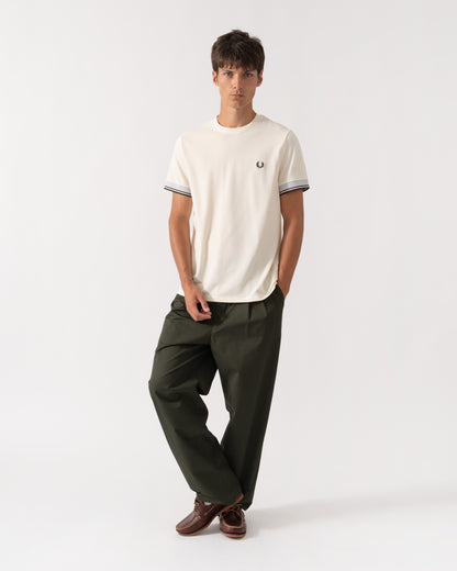 A man modeling a cream short-sleeve t-shirt with striped cuffs and a small embroidered logo, paired with dark olive green trousers and brown shoes, against a plain white studio background.
