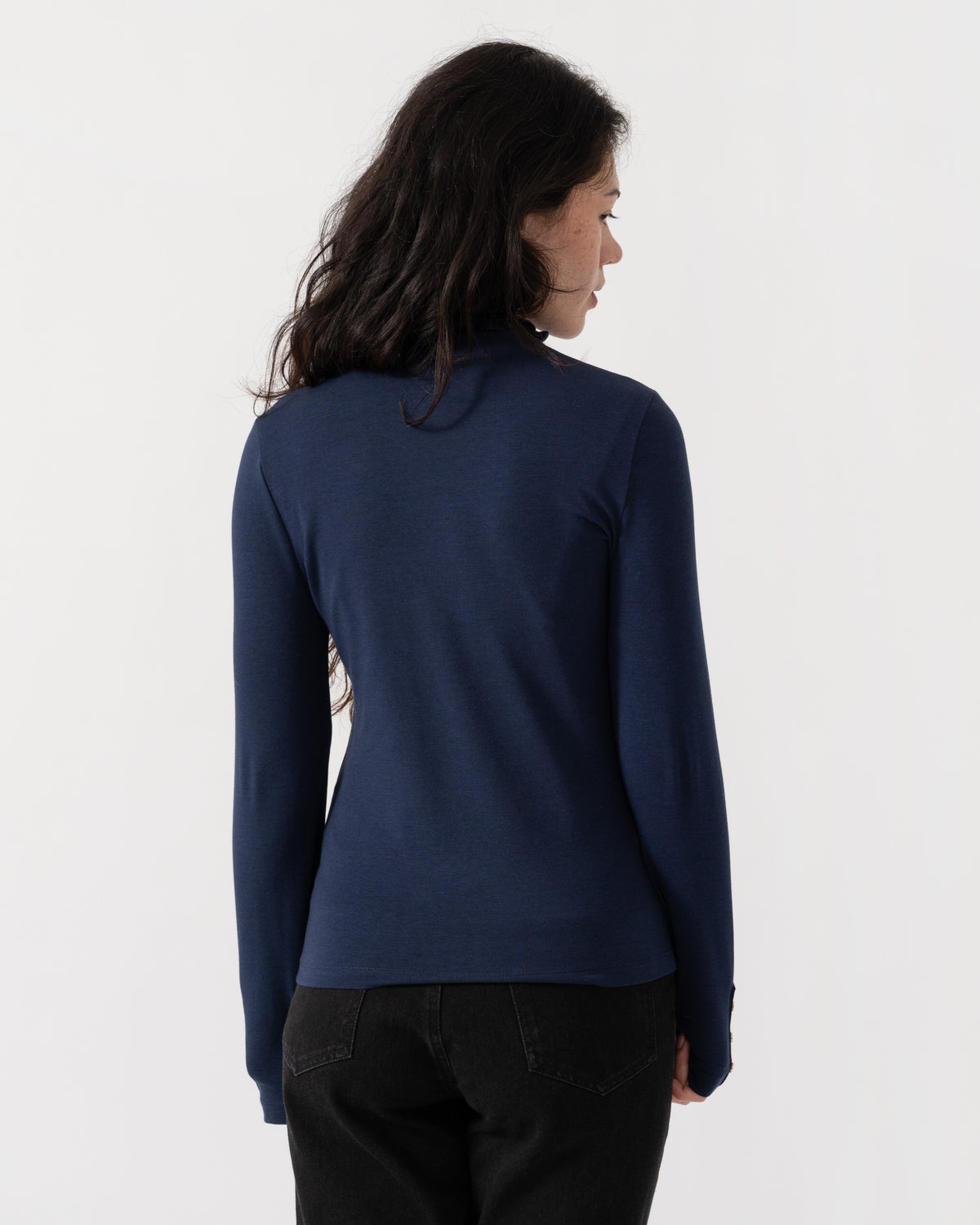 A woman with long dark hair is shown from the back wearing a navy blue long-sleeve top and black pants against a plain white studio background.