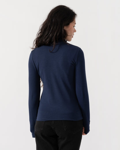 A woman with long dark hair is shown from the back wearing a navy blue long-sleeve top and black pants against a plain white studio background.