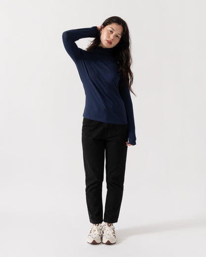 A woman modeling a dark navy blue long-sleeve turtleneck top, paired with black pants and white sneakers, against a plain white studio background.