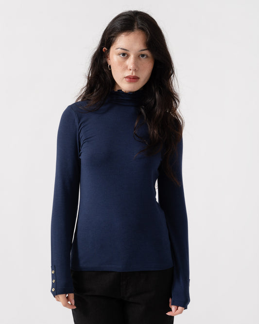 A woman modeling a navy blue long-sleeve mock neck top with decorative gold buttons on the cuffs, paired with black pants, against a plain light gray background.