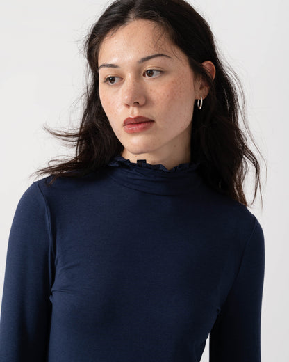 A woman wearing a dark navy blue long-sleeve mock neck top with a ruffled trim, photographed against a plain light gray background.