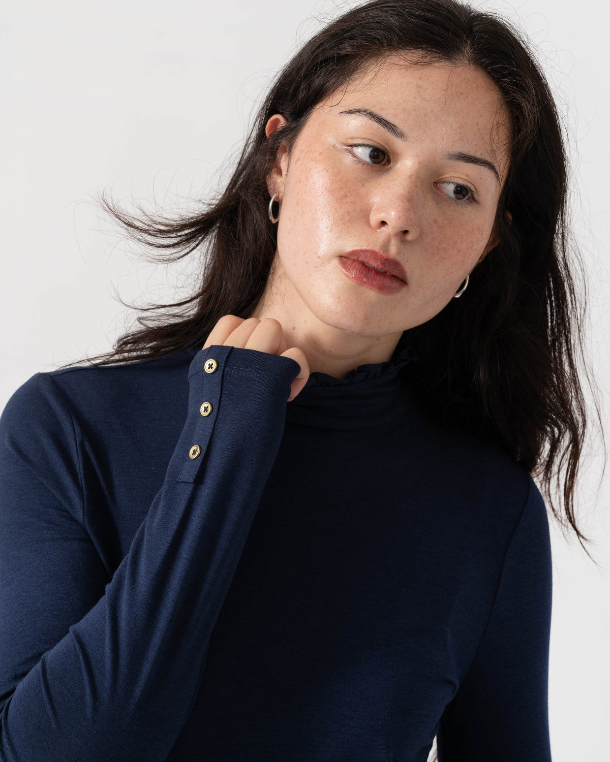 A woman wearing a navy blue long-sleeve mock neck top with gold buttons on the cuff, photographed against a clean light gray background.