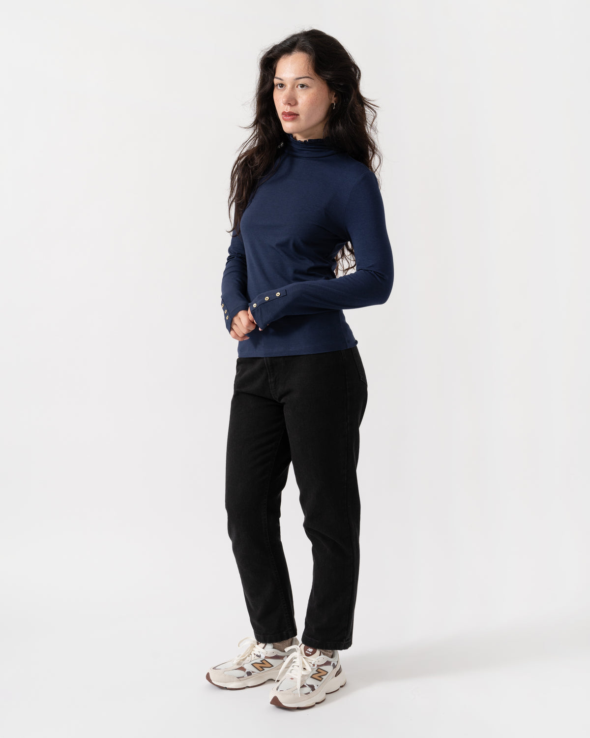 A woman modeling a navy blue mock neck long-sleeve top with patterned cuffs, paired with black pants and white sneakers, against a plain white studio background.