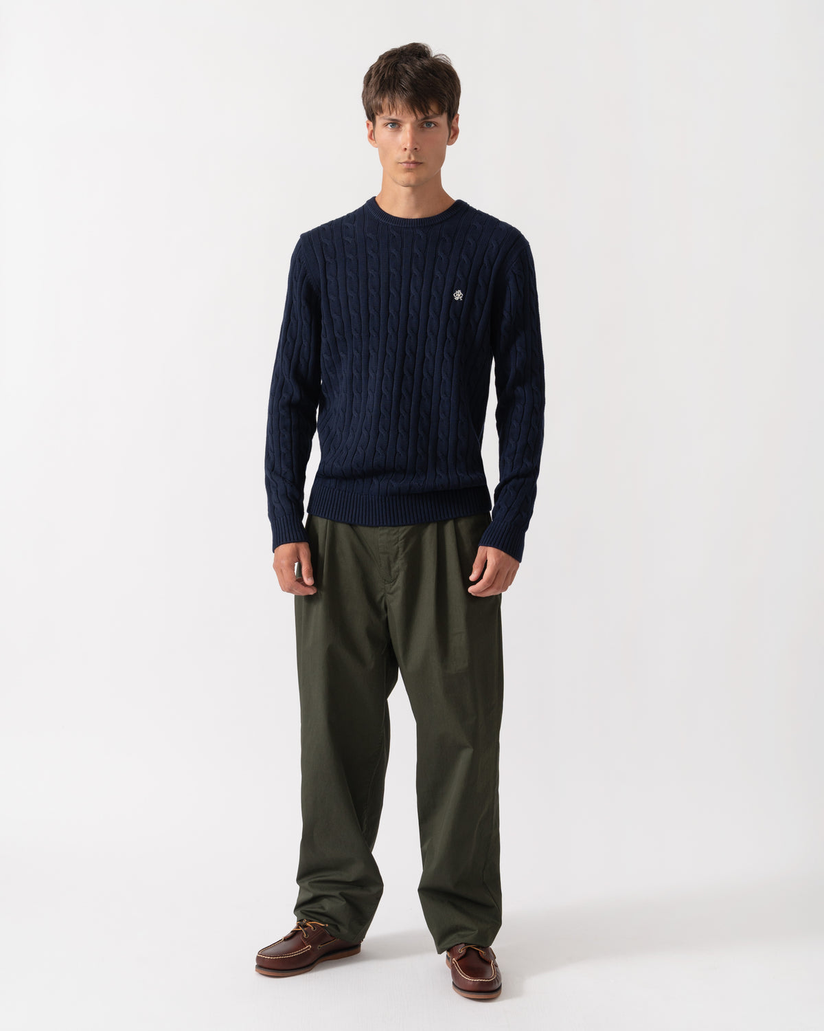 A man modeling a navy blue cable-knit crewneck sweater with a small white embroidered logo on the chest, paired with olive green trousers and brown leather shoes, against a plain white studio background.
