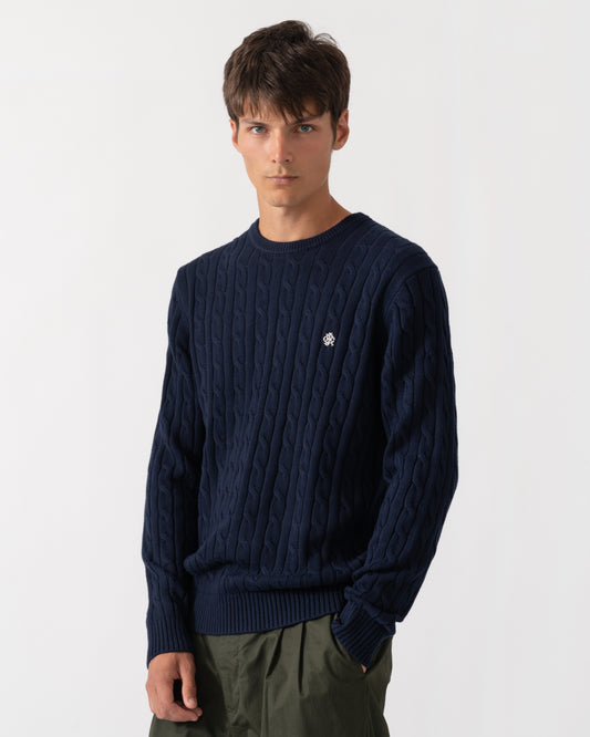 A man wearing a navy blue cable-knit crewneck sweater with a small embroidered logo on the chest, paired with olive green trousers, against a plain white studio background.