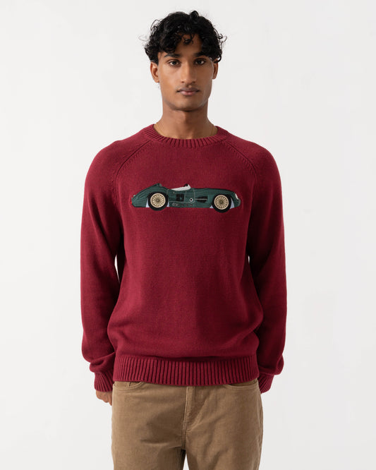 A man wearing a burgundy crewneck sweater with a green vintage race car graphic on the chest, paired with tan trousers, against a plain light gray background.