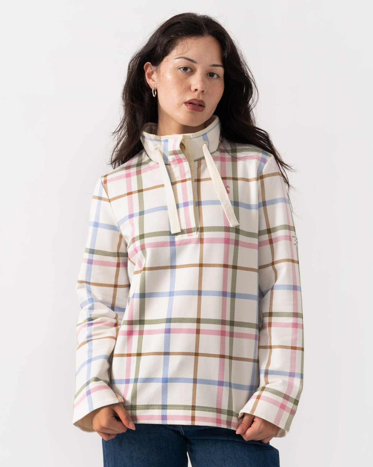 A woman wearing a cream-colored long-sleeve pullover with a pastel plaid pattern in pink, blue, green, and brown, paired with blue jeans, against a plain white studio background.