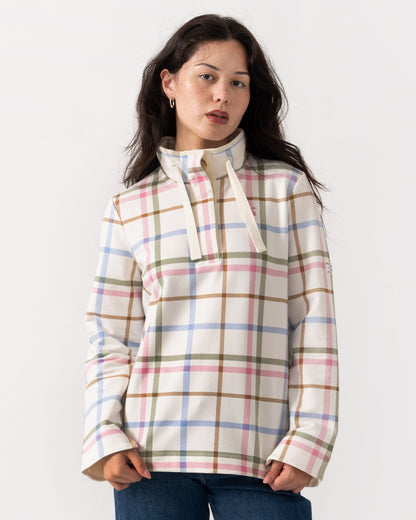 A woman wearing a cream-colored long-sleeve pullover with a pastel plaid pattern in pink, blue, green, and brown, paired with blue jeans, against a plain white studio background.