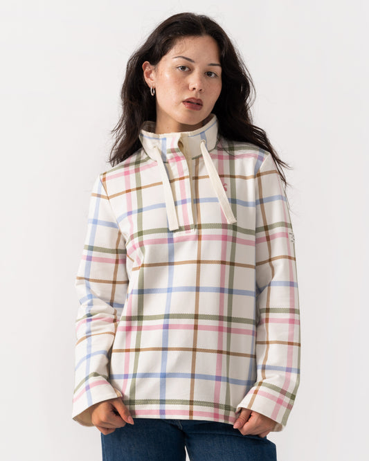 A woman wearing a cream-colored long-sleeve pullover with a pastel plaid pattern in pink, blue, green, and brown, paired with blue jeans, against a plain white studio background.