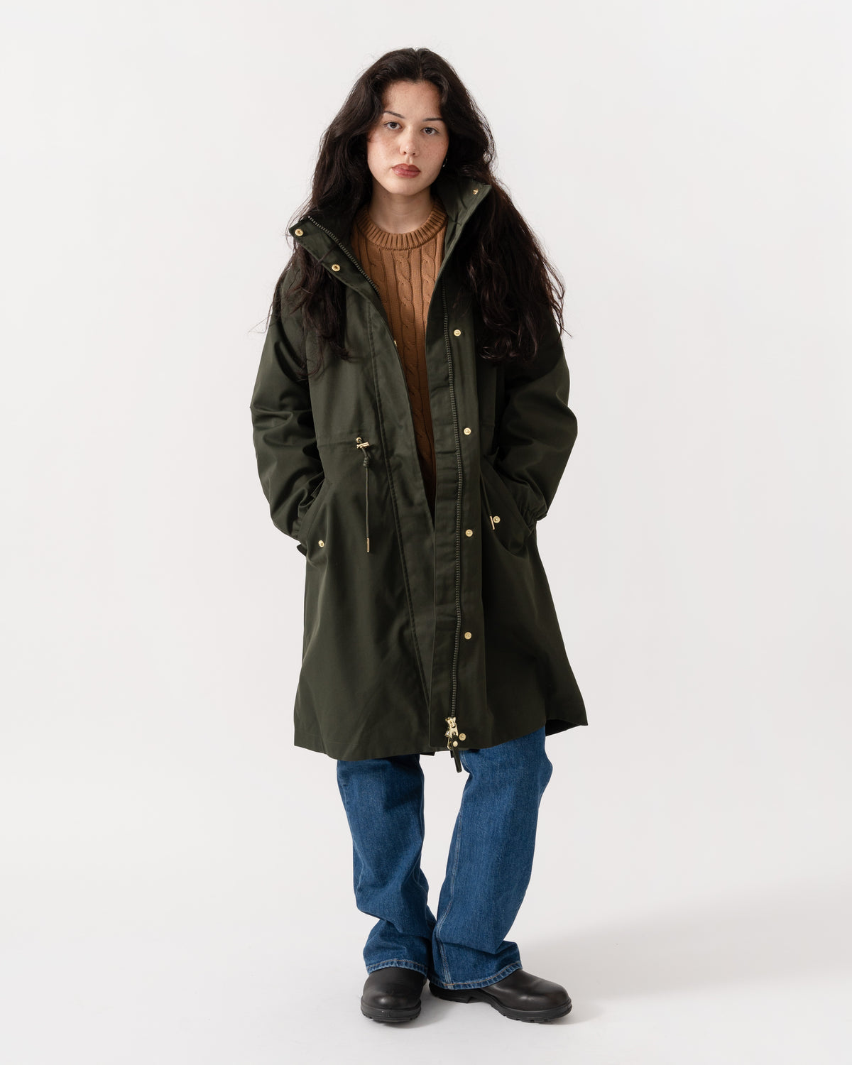 A woman modeling a dark olive green hooded parka jacket with gold-tone snap buttons, worn over a brown ribbed sweater and blue jeans, against a plain white studio background.