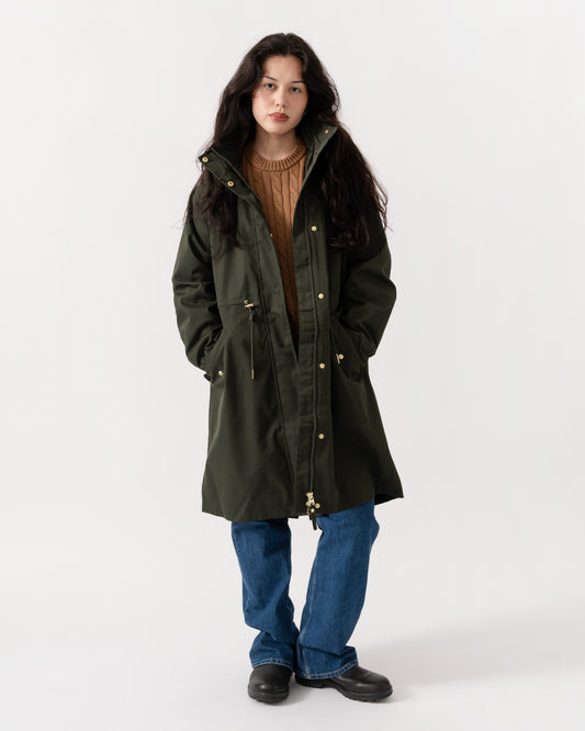 A woman modeling a dark olive green hooded parka jacket with gold-tone snap buttons, worn over a brown ribbed sweater and blue jeans, against a plain white studio background.