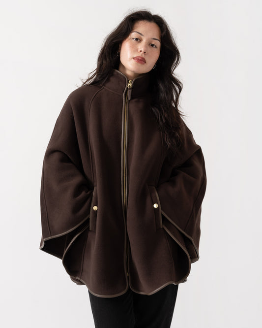 A woman wearing a dark brown cape-style jacket with a high collar, front zipper, and gold-tone snap details, photographed against a plain light gray studio background.
