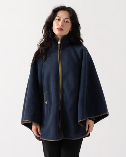 A woman wearing a navy blue cape-style coat with a high collar, front zipper, and wide draped sleeves, photographed against a plain light gray studio background.
