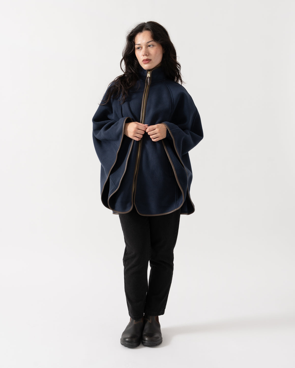 A woman modeling a navy blue poncho-style jacket with gold-toned zipper and piping details, paired with black pants and black boots, against a plain white studio background.