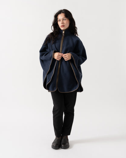 A woman modeling a navy blue poncho-style jacket with gold-toned zipper and piping details, paired with black pants and black boots, against a plain white studio background.
