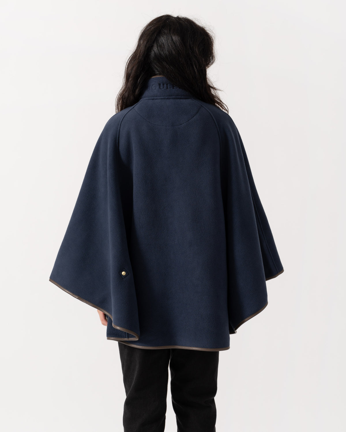 A person shown from the back wearing a navy blue cape-style coat with wide draped sleeves and a small gold button detail near the lower left hem, against a plain light background.