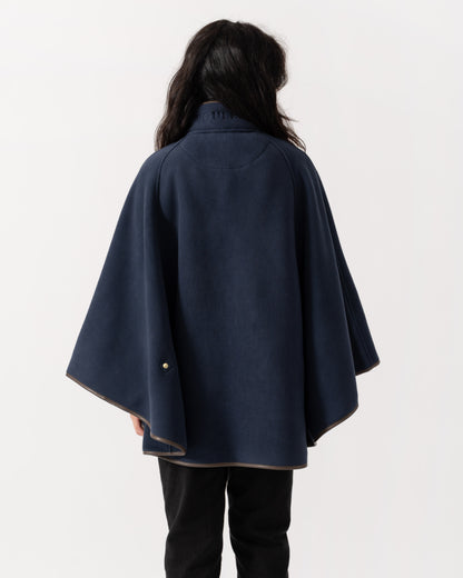 A person shown from the back wearing a navy blue cape-style coat with wide draped sleeves and a small gold button detail near the lower left hem, against a plain light background.