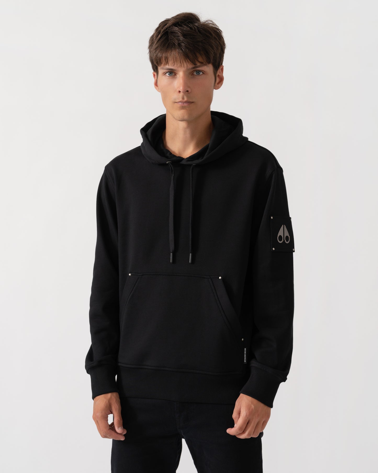 A man wearing a black pullover hoodie with drawstrings, a kangaroo pocket, and a small white embroidered logo on the left sleeve, photographed against a light gray studio background.