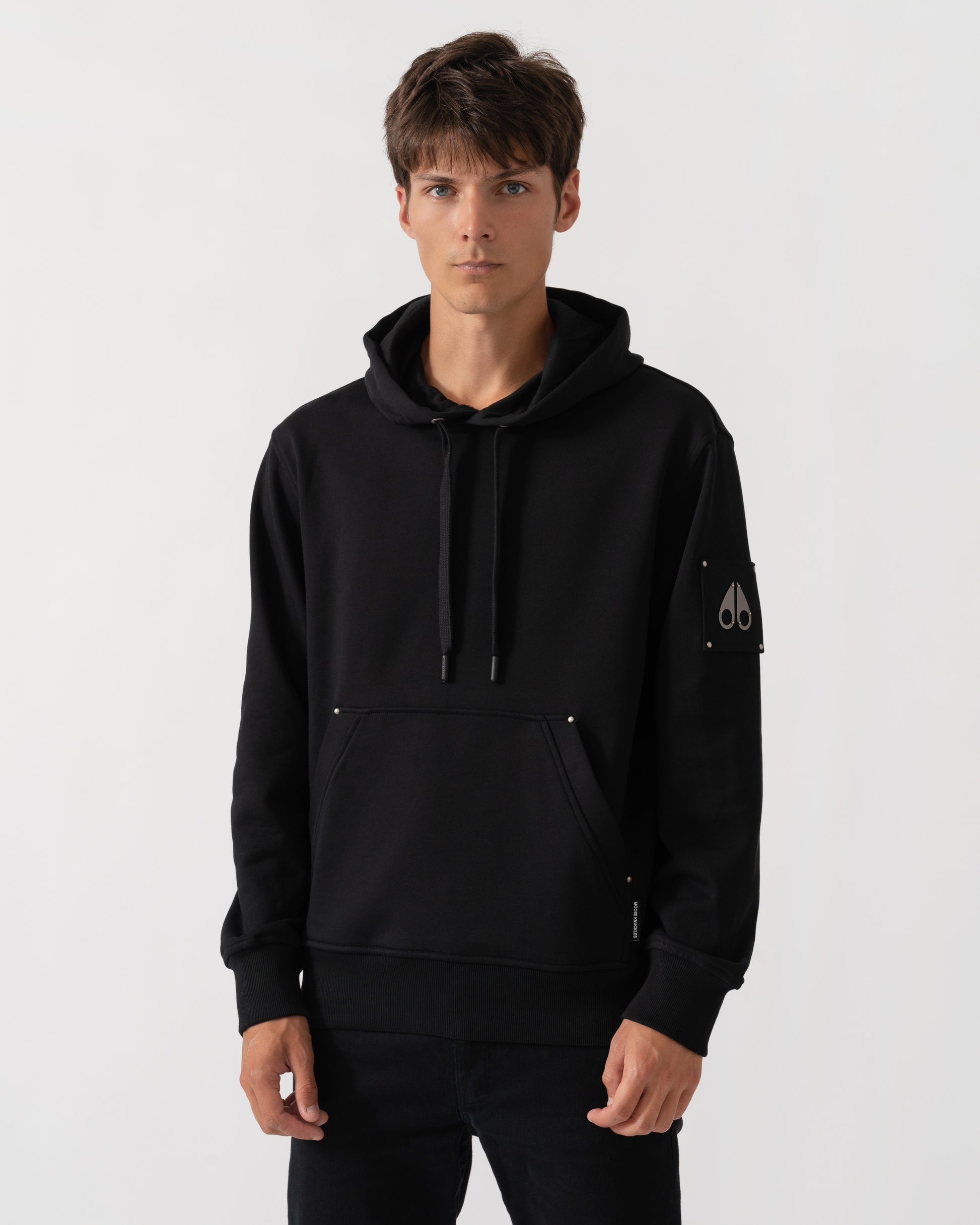 A man wearing a black pullover hoodie with drawstrings, a kangaroo pocket, and a small white embroidered logo on the left sleeve, photographed against a light gray studio background.