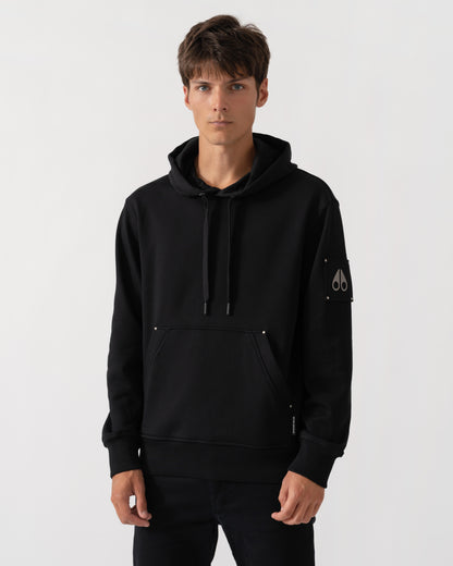 A man wearing a black pullover hoodie with drawstrings, a kangaroo pocket, and a small white embroidered logo on the left sleeve, photographed against a light gray studio background.