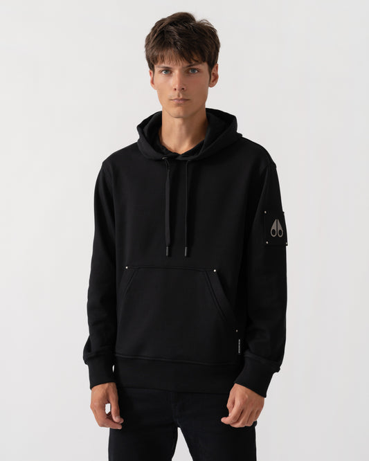 A man wearing a black pullover hoodie with drawstrings, a kangaroo pocket, and a small white embroidered logo on the left sleeve, photographed against a light gray studio background.