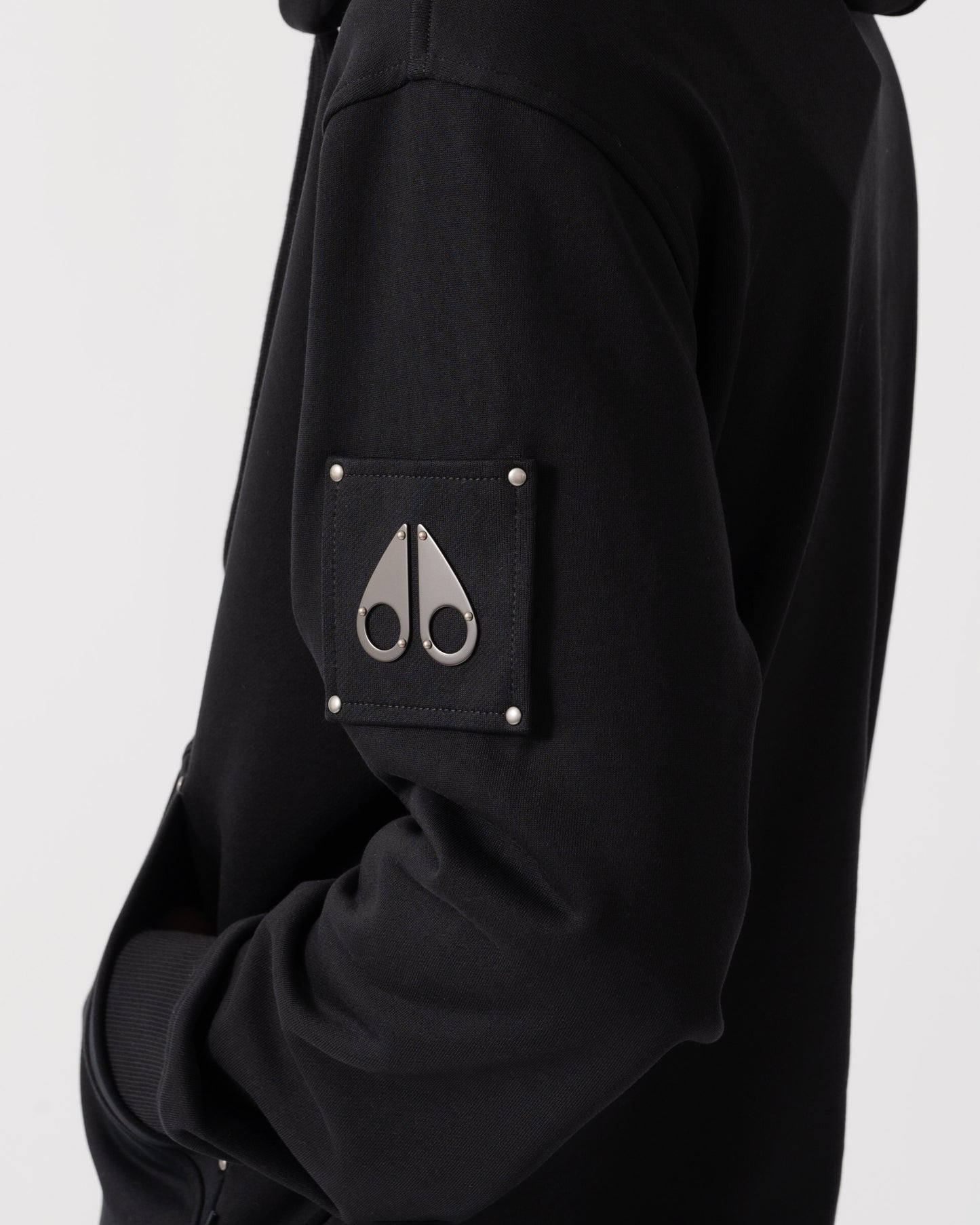 Close-up of a black hooded jacket featuring a square chest pocket with silver rivets and a silver metal scissors logo on the front.