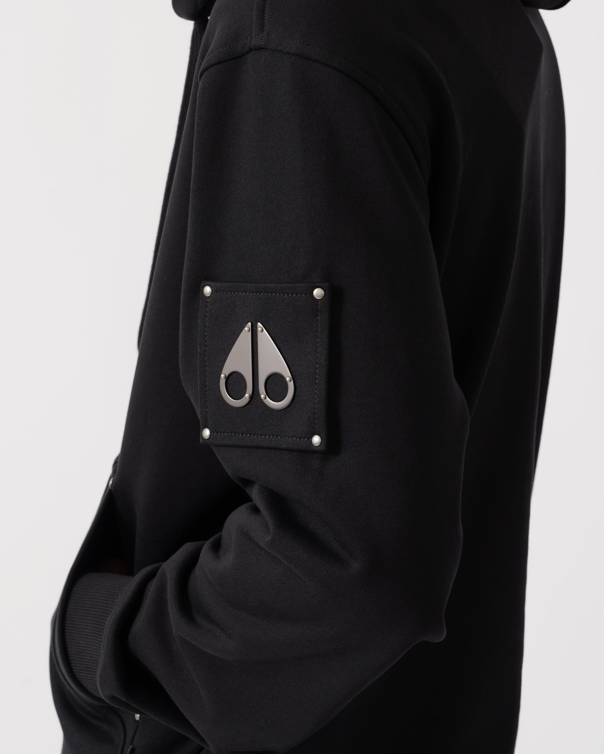 Close-up of a black hooded jacket featuring a square chest pocket with silver rivets and a silver metal scissors logo on the front.