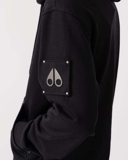 Close-up of a black hooded jacket featuring a square chest pocket with silver rivets and a silver metal scissors logo on the front.