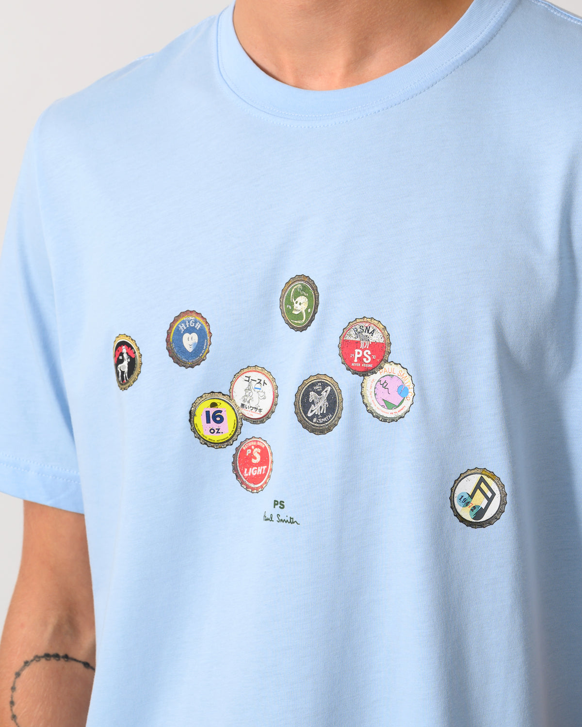 close up studio photograph of a model wearing a PS Paul Smith Mens Regular Fit Bottle Tops Graphic T-Shirt in 41E Light Blue