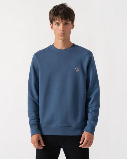 A man wearing a slate blue crewneck sweatshirt with a small embroidered dog logo on the left chest, paired with black pants, against a plain light gray background.