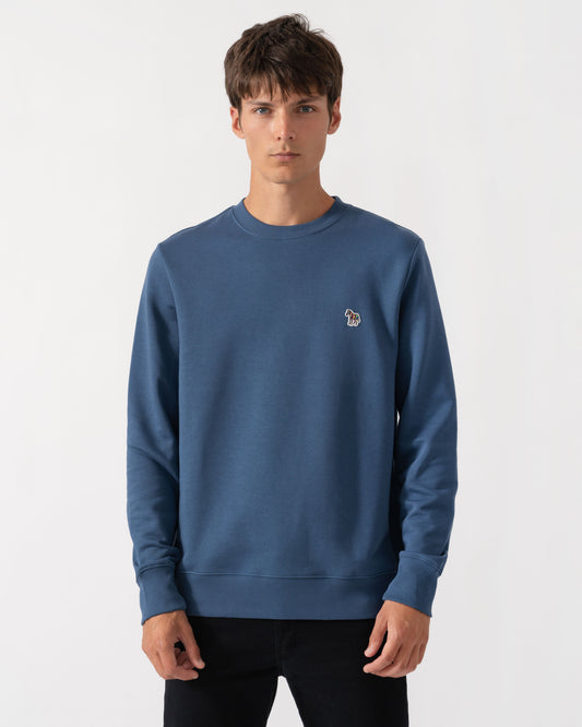 A man wearing a slate blue crewneck sweatshirt with a small embroidered dog logo on the left chest, paired with black pants, against a plain light gray background.