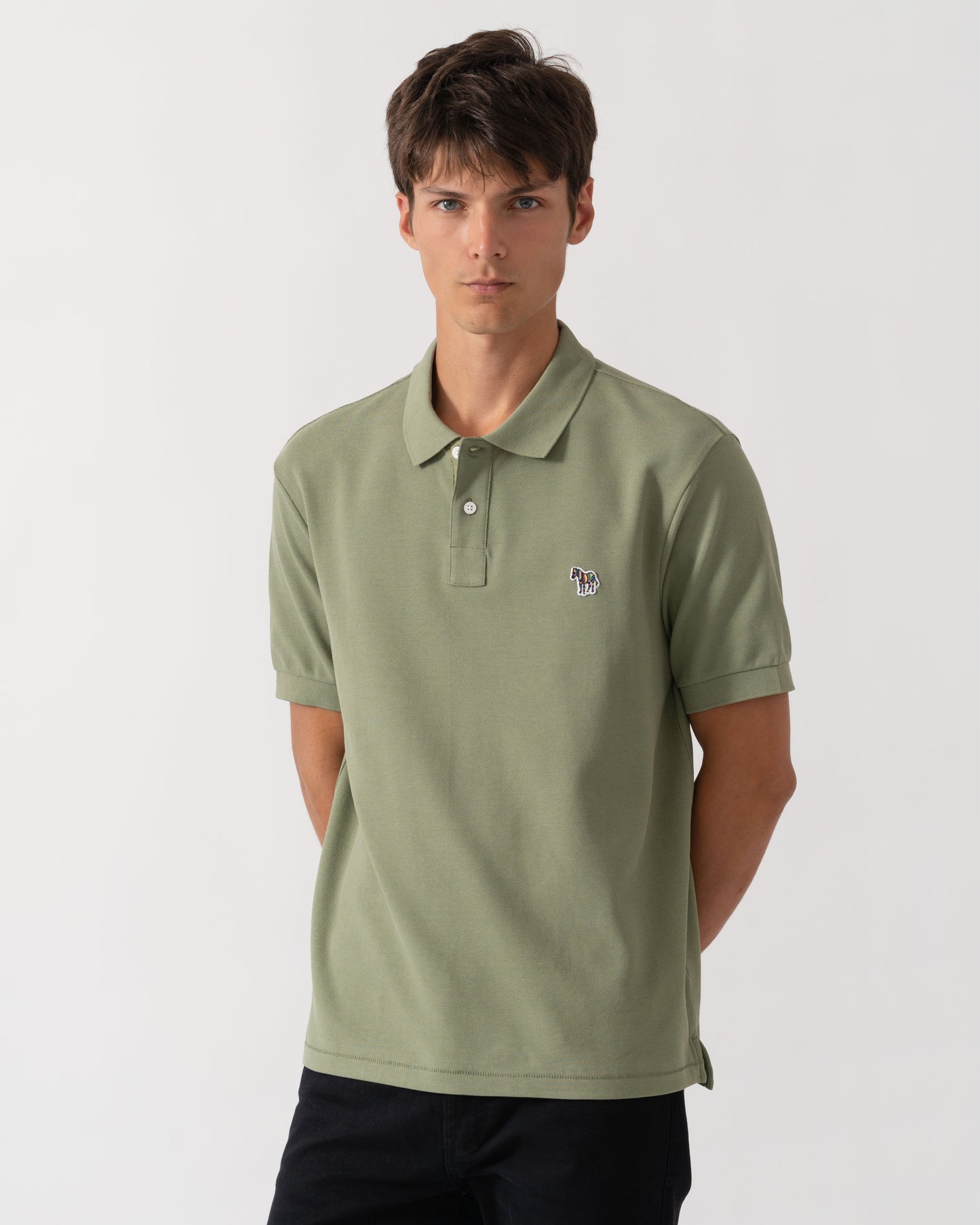 A man wearing a sage green short-sleeve polo shirt with a small embroidered logo on the left chest, paired with dark pants, photographed against a light gray studio background.