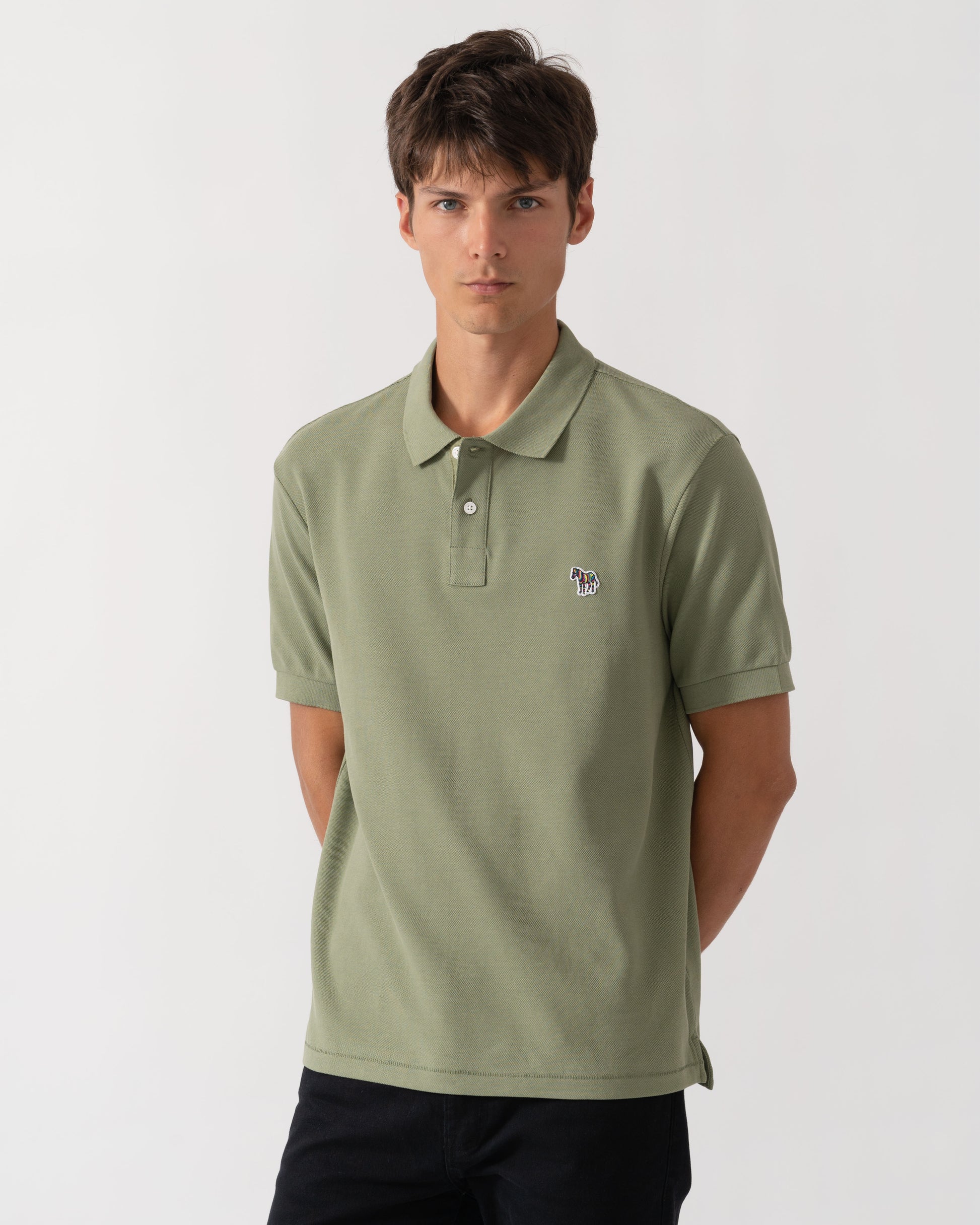 A man wearing a sage green short-sleeve polo shirt with a small embroidered logo on the left chest, paired with dark pants, photographed against a light gray studio background.
