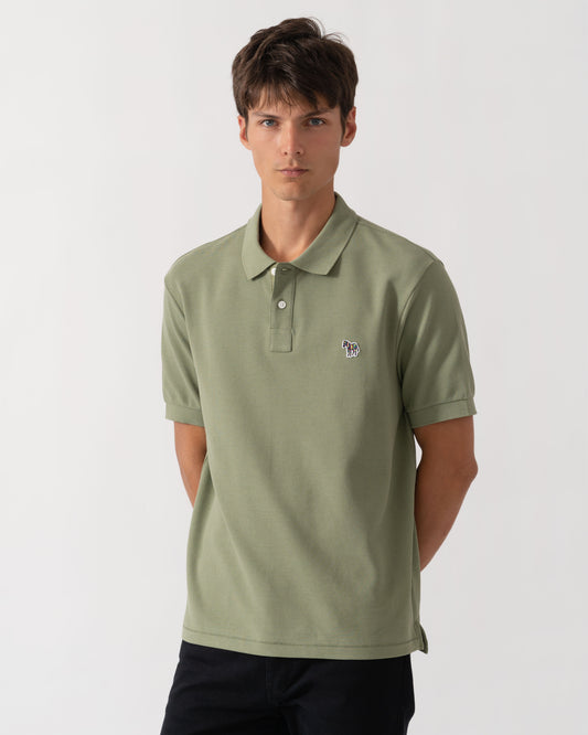 A man wearing a sage green short-sleeve polo shirt with a small embroidered logo on the left chest, paired with dark pants, photographed against a light gray studio background.
