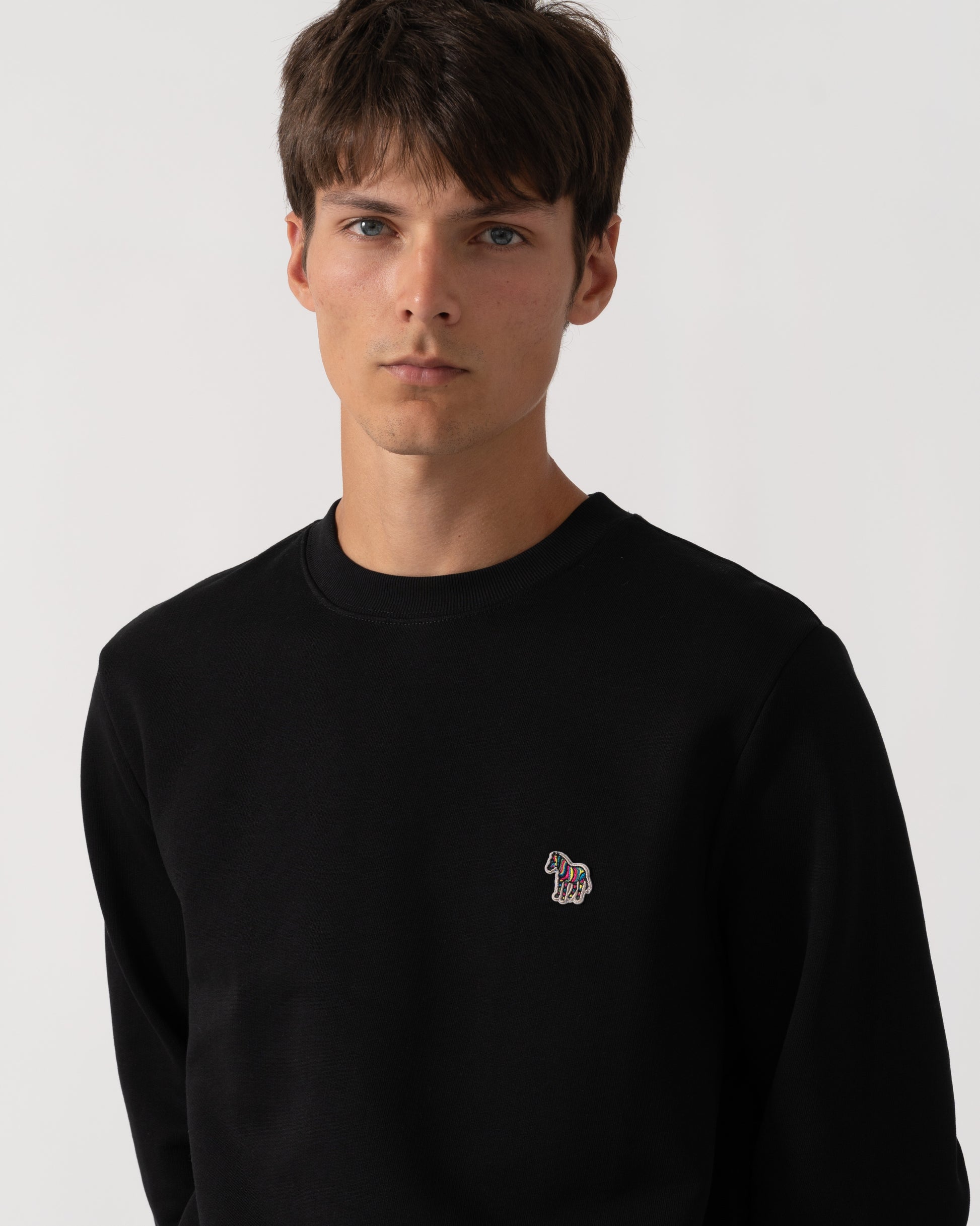 A man wearing a black crewneck sweatshirt with a small embroidered horse logo on the left chest, photographed against a light gray studio background.
