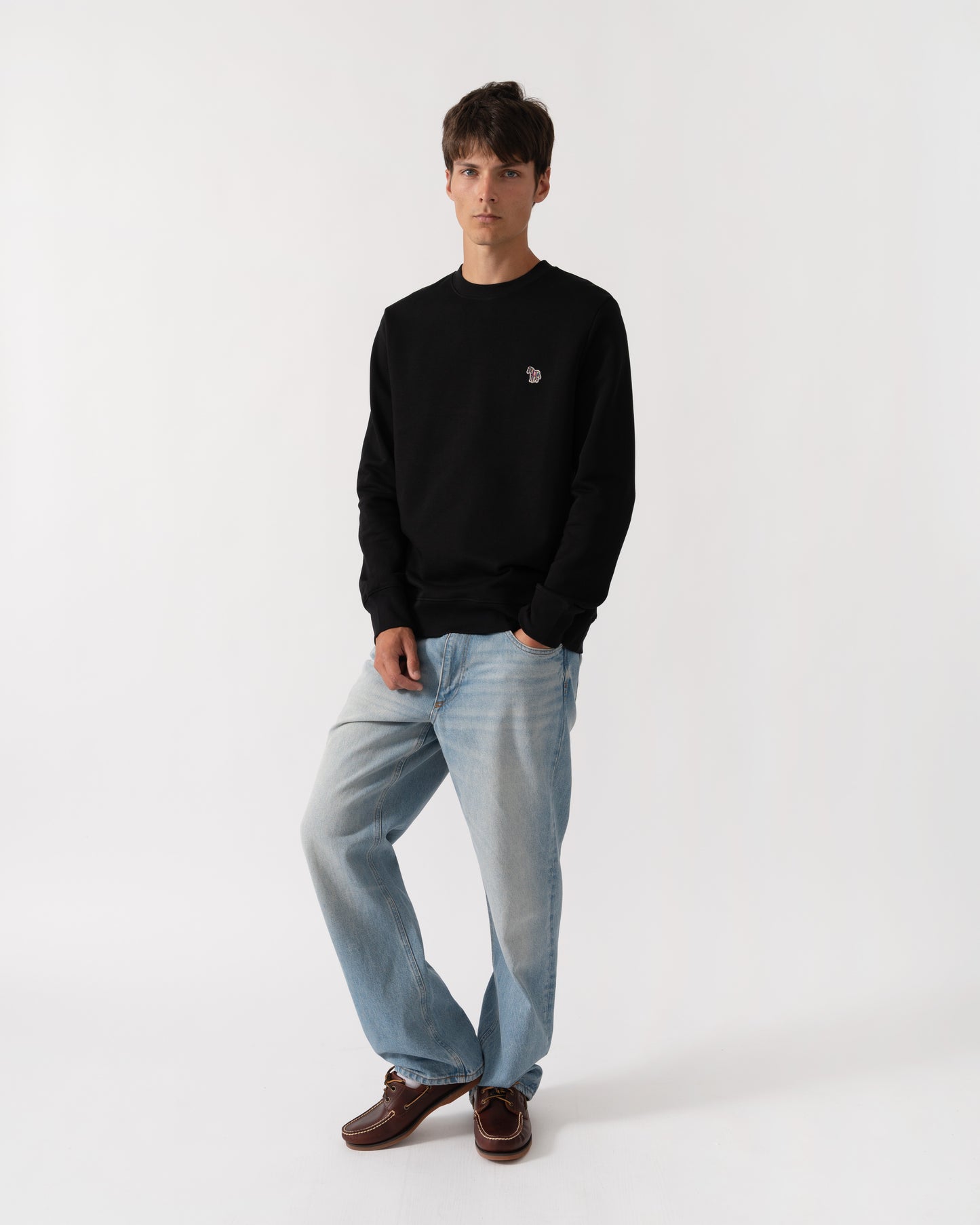 A man modeling a black crewneck sweatshirt with a small embroidered logo on the left chest, paired with light blue jeans and brown leather shoes, against a plain white studio background.