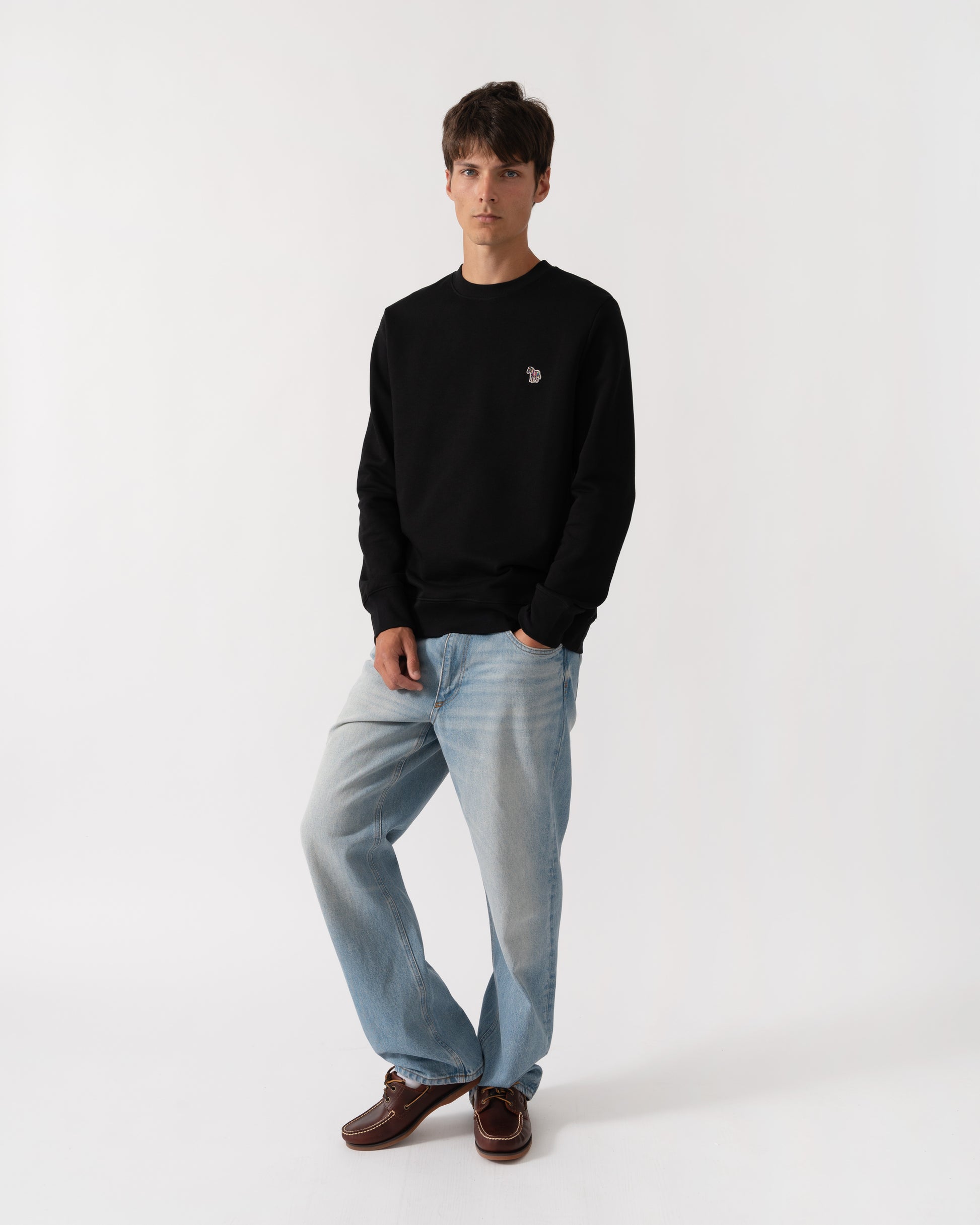 A man modeling a black crewneck sweatshirt with a small embroidered logo on the left chest, paired with light blue jeans and brown leather shoes, against a plain white studio background.